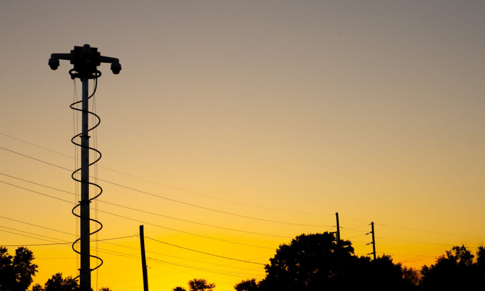 Silhouetted security camera tower at sunset with trees nearby.