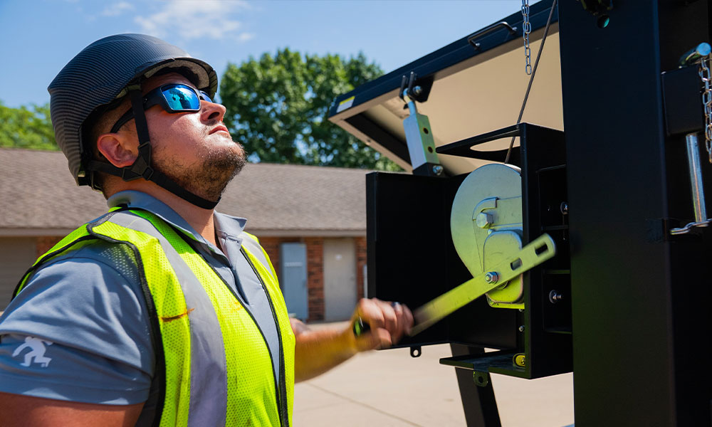 Man in safety gear operates crank handle outdoors near building.