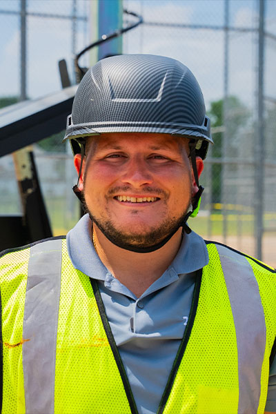 Man in black hard hat and yellow vest smiles outdoors.
