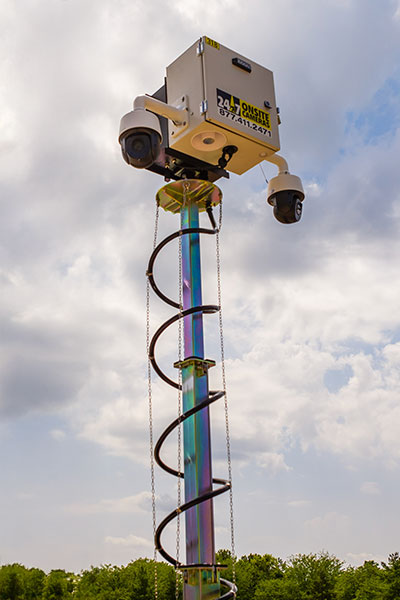 Surveillance camera on spiral pole, cloudy sky and trees.