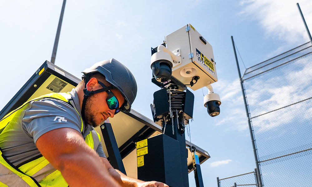 Worker adjusts equipment by surveillance tower under blue sky.