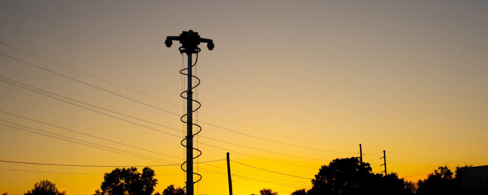 Silhouetted security camera tower at sunset with trees nearby.